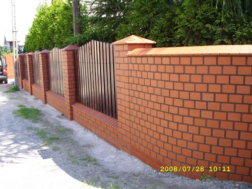 Ogrodzenie z cegły klinkierowej z drewnianymi przęsłami, zwieńczone daszkami z cegły, widok z zewnątrz na tle zieleni.